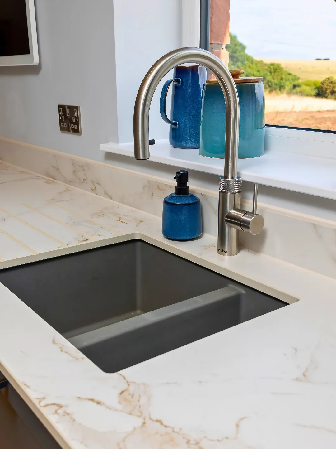 Kitchen tap, inset sink and pale stone worktop with blue glassware by the window